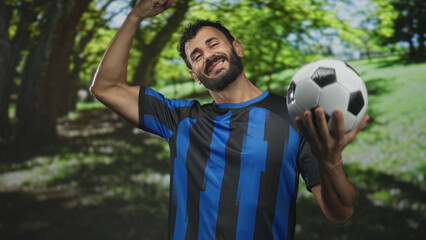 Obraz premium Man bearded soccer player in blue uniform holds soccer ball and flexes bicep while smiling in forest; pride.