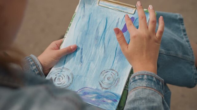 Artist painting blue watercolor on clipboard, denim jacket on lap, sitting on park bench with soft sunlight, gentle finger strokes creating wave textures and circular details, calm meditative