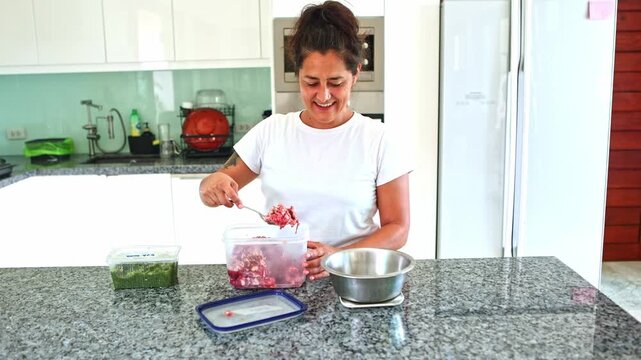 Smiling woman preparing BARF raw dog food in her modern kitchen