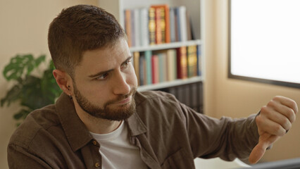 Obraz premium Man at computer shows thumbs down with right hand beside monitor in building, bookshelf and plant visible; frustration concentration.