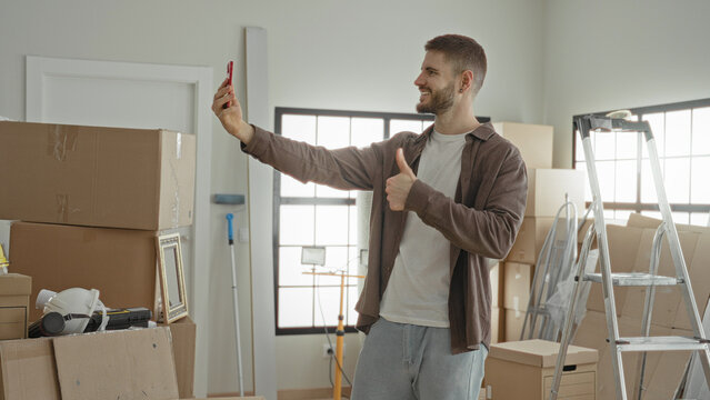 Man takes selfie with smartphone amid packed moving boxes, ladder, helmet and bubblewrap in building while unpacking and organizing; optimism new beginning.