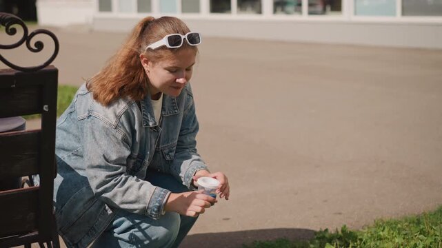 Teen girl outside holding pillbox by bench, denim jacket and sunglasses, modern building backdrop, careful hands inspecting medication container, candid campus lifestyle, warm sunlight, calm