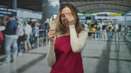 Fototapeta premium Woman holding makeup brushes, covering one eye and wearing red apron in airport check in line; travel fatigue.