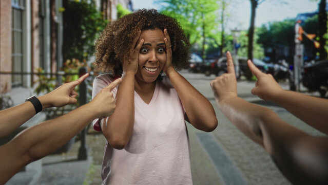 Woman with hands on head and multiple fingers pointing at her on a busy city street with parked cars; isolation anxiety.