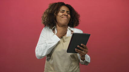 Woman baker in apron clutching neck and holding tablet in pink studio, eyes closed and face grimacing  pain stress. © Krakenimages.com