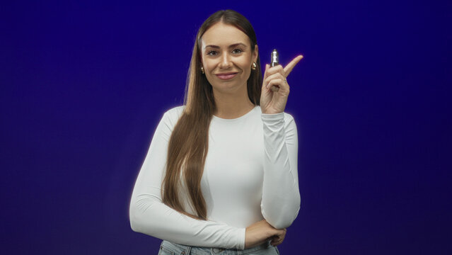 Woman holding a vaporizer while pointing her index finger, smiling and wearing a white top in studio; confidence composure.