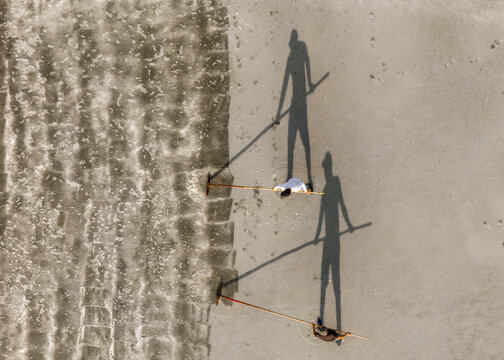 Aerial view of two workers raking salt in a field with long shadows cast on the textured ground in Dong Hai, Ninh Thuan, Vietnam.