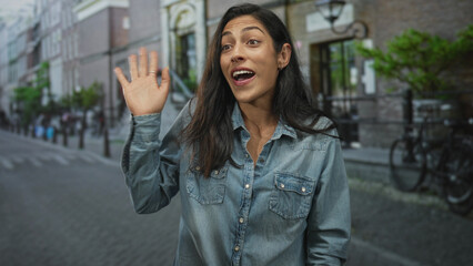Fototapeta premium Woman smiling with face visible and waving hand on a cobblestone street by a building; friendly joy.