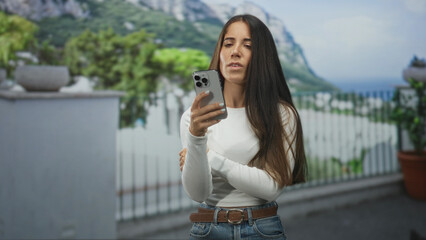 Fototapeta premium Woman holds smartphone with arms crossed on building balcony while checking the screen, neutral expression and relaxed posture; contemplation.