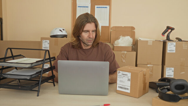 Man holding cardboard parcel while using laptop at desk amid stacked boxes in a shipping office building; focused work.