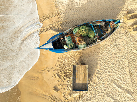 Aerial view of a traditional blue fishing boat filled with colorful nets on the sandy shore with tire tracks and ocean waves at Praia de Mira, Coimbra, Portugal.