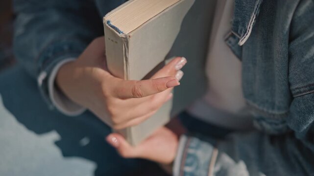Young woman holding book on lap, denim jacket and manicured nails bathed in warm sunlight closeup shows textured hardcover, gentle grip, relaxed posture on outdoor bench, calm weekend study moment