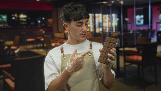 Man points finger to chocolate bar in building while holding bar with gloved hand; tasting curious review.