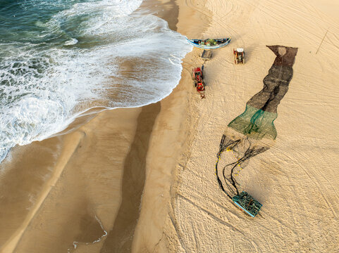 Aerial view of traditional fishing boats and nets on the sandy shore with tractors and crashing waves at Praia de Mira, Coimbra, Portugal.