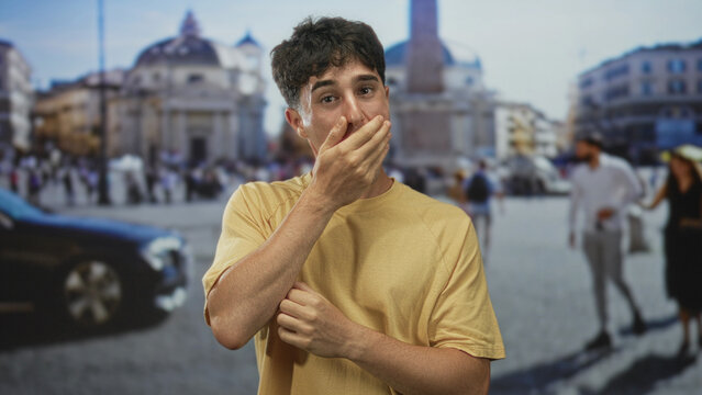 Young hispanic man covers mouth with hands standing outdoors in front of large church building at st peter square, vatican; surprise secrecy.