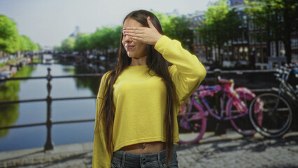 Obraz premium Teen girl in yellow sweater covers eyes with hand, exposed navel and long hair, standing by canal railing with parked bicycles on street; playful.