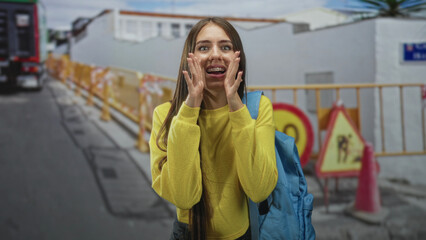 Fototapeta premium Teenage girl with blue backpack clenches fists near face, smiling with braces in front of street construction barriers and cones; joy celebration.