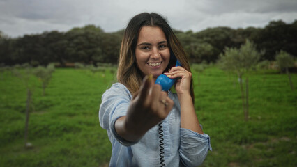 Fototapeta premium Woman smiling and pointing finger at camera while holding a blue handset amid a green forest field with trees and grass; playful connection.