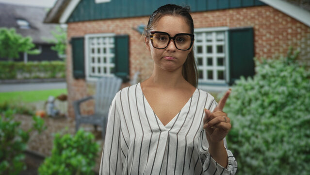 Woman in striped blouse wearing large glasses points finger to the right at a house building entrance outdoors; disapproval judgment.