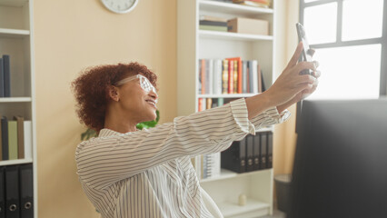 Woman holding smartphone smiling and raising arm to take selfie at desk with computer and bookshelves in building  workday joy. © Krakenimages.com