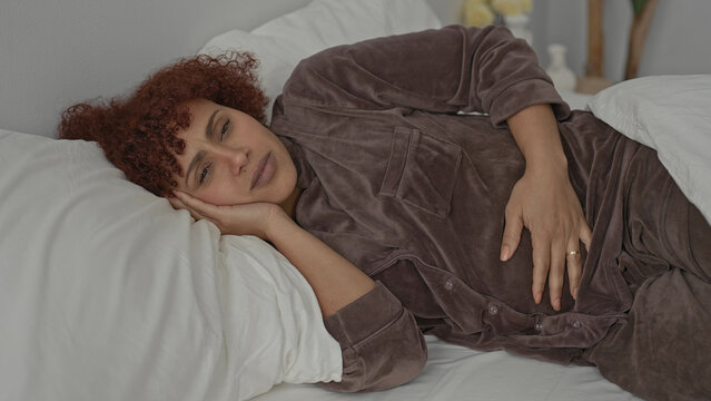 Hispanic brunette woman clutching stomach with hand to head on white bed in studio, pained expression wearing brown pajamas for stomachache; discomfort.