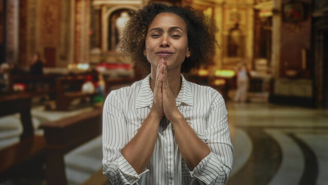 Woman praying with hands clasped inside a church building, eyes closed and head tilted upward in worship; devotion.
