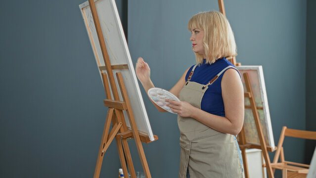 Woman blonde painter wearing apron and blue top, painting on canvas at wooden easel with brush and palette, hand poised while standing in studio; serene focus.