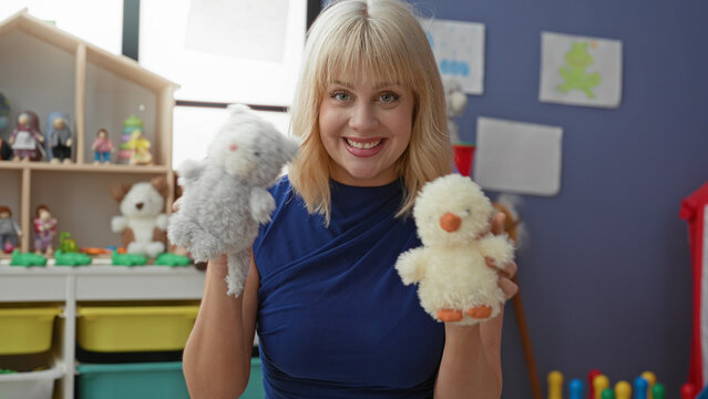 Woman holds two plush toys up with hands while smiling directly at camera in a colorful preschool building play area, engaging in puppet play; teaching play joy.