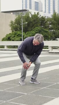 Vertical video: Walking man with knee pain, holding knee and limping across tiled plaza in gray top