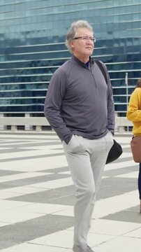 Vertical video: Walking man crossing plaza, camera tracking, fixing bag, woman on phone with coffee