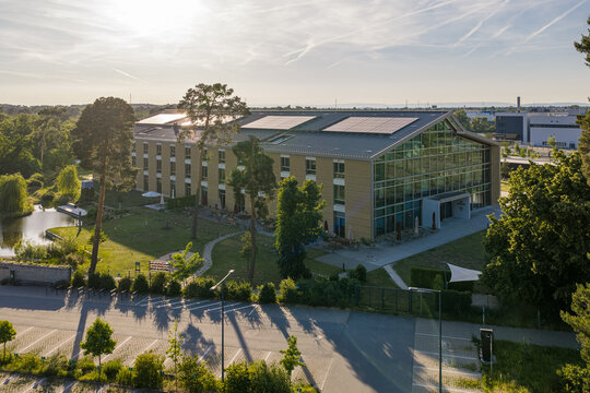 Darmstadt, Germany - 25 May 2026: Aerial view of the Alnatura Campus building with solar panels on the roof, surrounded by green trees and a small pond under a bright afternoon sun.