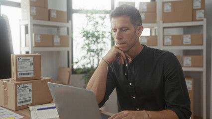 Fototapeta premium Man working on laptop with hand on chin in office building, surrounded by stacked shipping boxes and shelving; thoughtful planning.