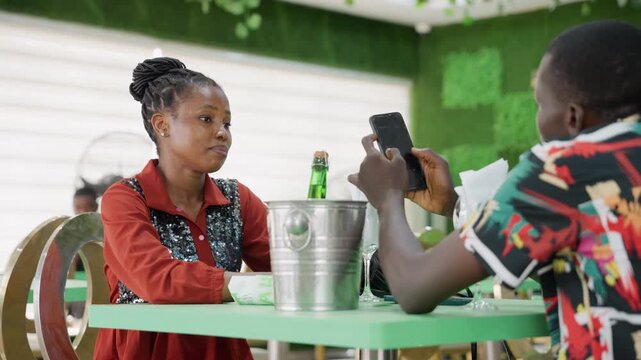 Awkward cafe date with black woman. Woman in red jacket watches partner show phone across table, ice bucket and green bottle between them, subtle tension and distracted gaze, scrolling