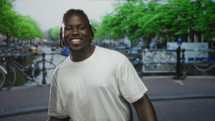 Fototapeta premium Man smiling showing teeth on street beside an amsterdam canal with bicycles and railing; urban summer joy.