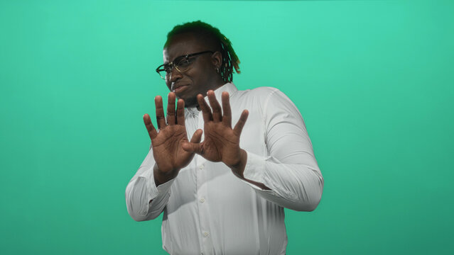 Man in white shirt showing palms out hands in studio with teal green backdrop, wearing glasses and dreadlocks; hesitation.