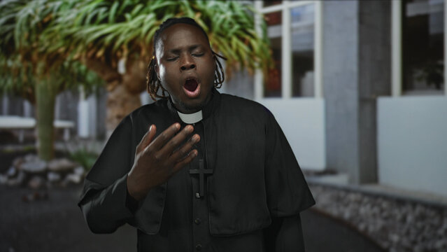 Man priest yawning with hand to mouth showing clerical collar and cross near building courtyard; tired contemplation.