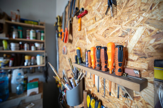 A row of screwdrivers with orange and black handles is organized on a wooden shelf attached to an oriented strand board wall inside a home workshop with storage shelves in the background