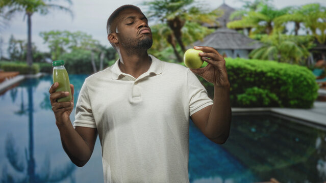 Man holding a green apple and juice bottle in bare hands beside a pool and resort building while examining the fruit; indecision healthy choice.