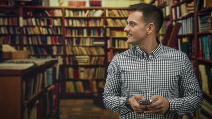 Man with moustache holding smartphone in hands and looking to the side in a building filled with bookshelves and rows of books  quiet curiosity. © Krakenimages.com