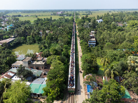 Akkelpur, Bangladesh - 05 June 2025: Aerial view of a train crowded with passengers on the roof moving along a straight track through lush green fields and rural settlements.