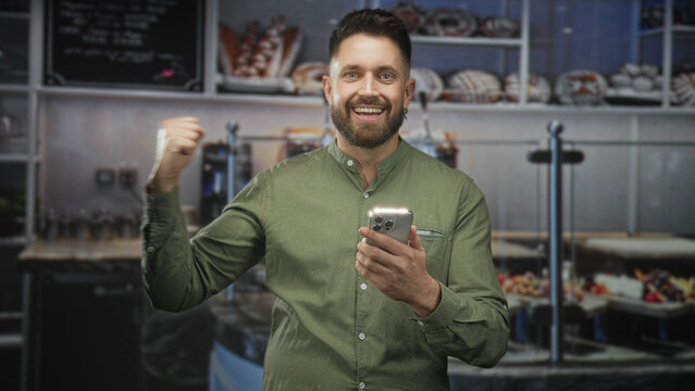 Hispanic bearded man holding smartphone clenches fist and laughs while looking up in bakery building near pastry counter; success win joy.