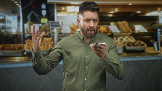 Man holding smartphone, hand gesturing and speaking into phone at bakery counter while disputing order with staff; frustration complaint.