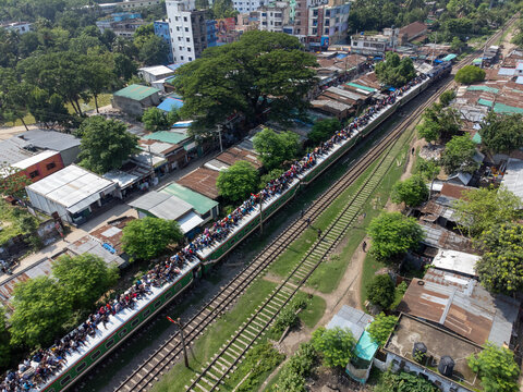 Akkelpur, Bangladesh - 05 June 2025: Aerial view of a crowded passenger train with people sitting on the roof traveling through the town surrounded by buildings and green trees.