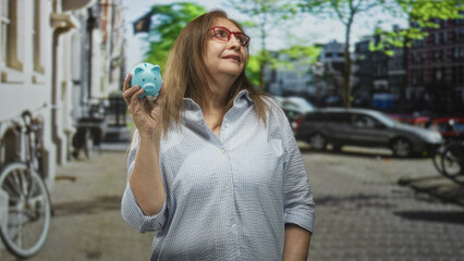 Fototapeta premium Senior hispanic woman wearing glasses holding a blue piggy bank in her hand on a street with parked cars and bicycle; financial planning hopeful.