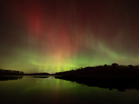 Northern lights of red and green fill the sky and reflect in the waters of a lake below