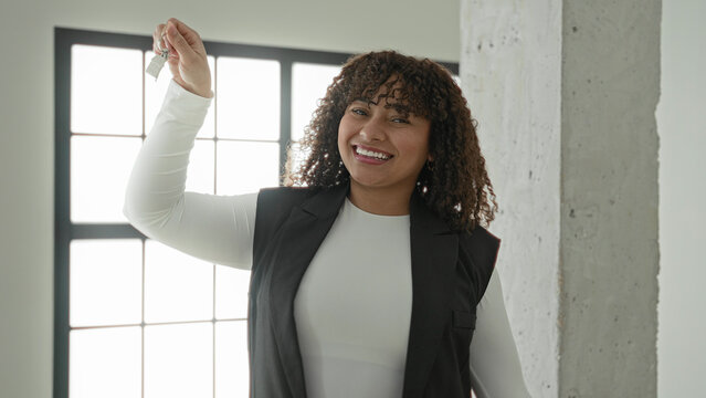 Woman with curly brunette hair holds keys high with hand raised, smiling in empty building interior by window and concrete pillar; new home joy.