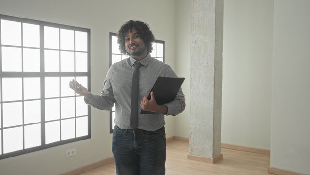 Man holding black folder gestures with open hand and points with other hand in empty building interior, smiling and presenting the space; confidence trust.