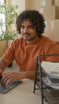 Man typing on laptop amid stacked cardboard boxes in an office building with parcel supplies; focused productivity.