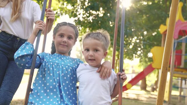 two children on a swing. concept of a happy childhood and loving family lifestyle. girl and boy in beautiful clothes swings on a swing on the playground, they are being filmed by their parents