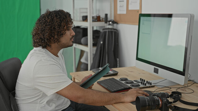 Man photographer taps tablet beside camera and desktop computer in studio at wooden desk with monitor and keyboard; focused creativity.